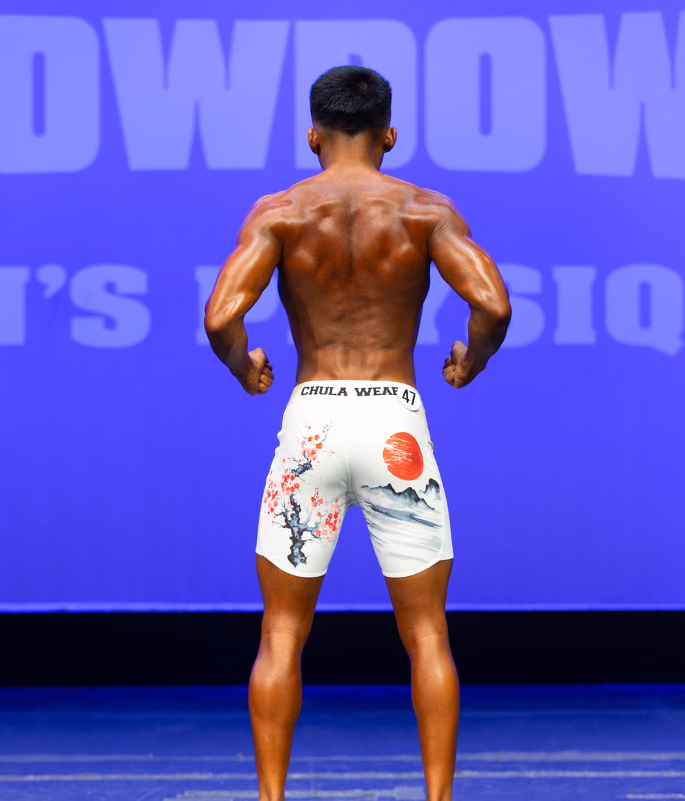 Mark Villanueva Jr. on stage at a men's physique competition, back pose